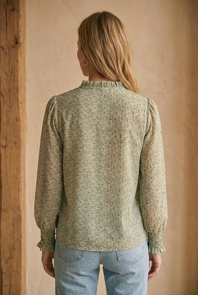 A woman viewed from the back wearing a long-sleeve blouse with a dense small floral print in muted green and beige, paired with light blue jeans, set against a warm textured beige wall.