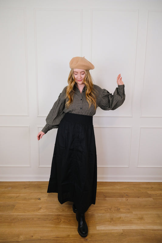 White woman in black maxi skirt and puffy long sleeve check button down shirt and a tan french beret