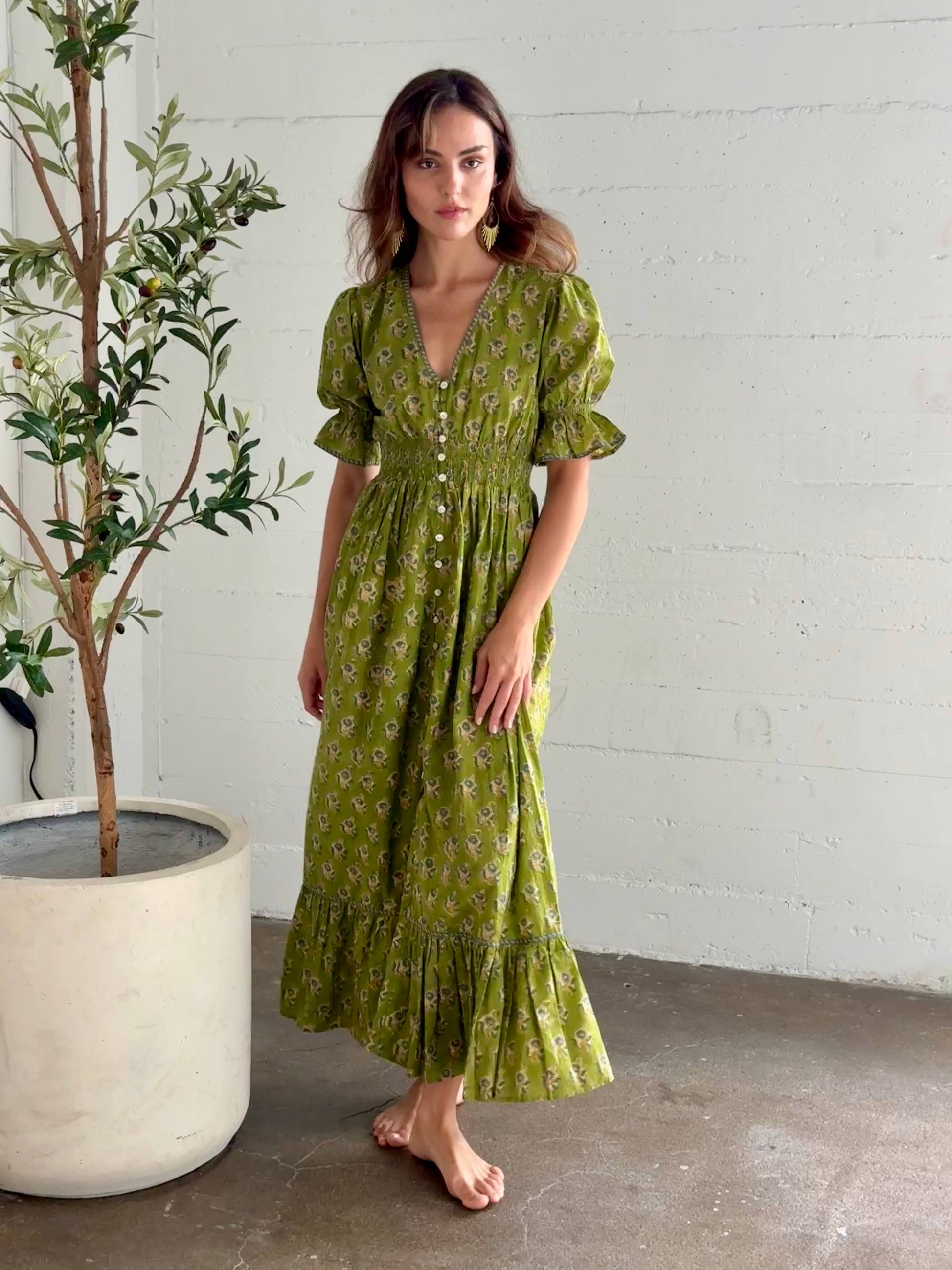 Woman wearing a green floral dress standing next to a potted plant against a white wall.