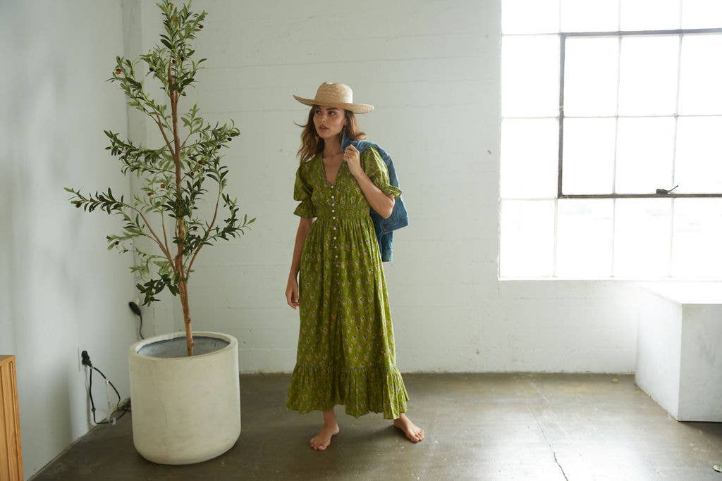 Woman in a green dress and hat standing next to a potted plant in a room with large windows.