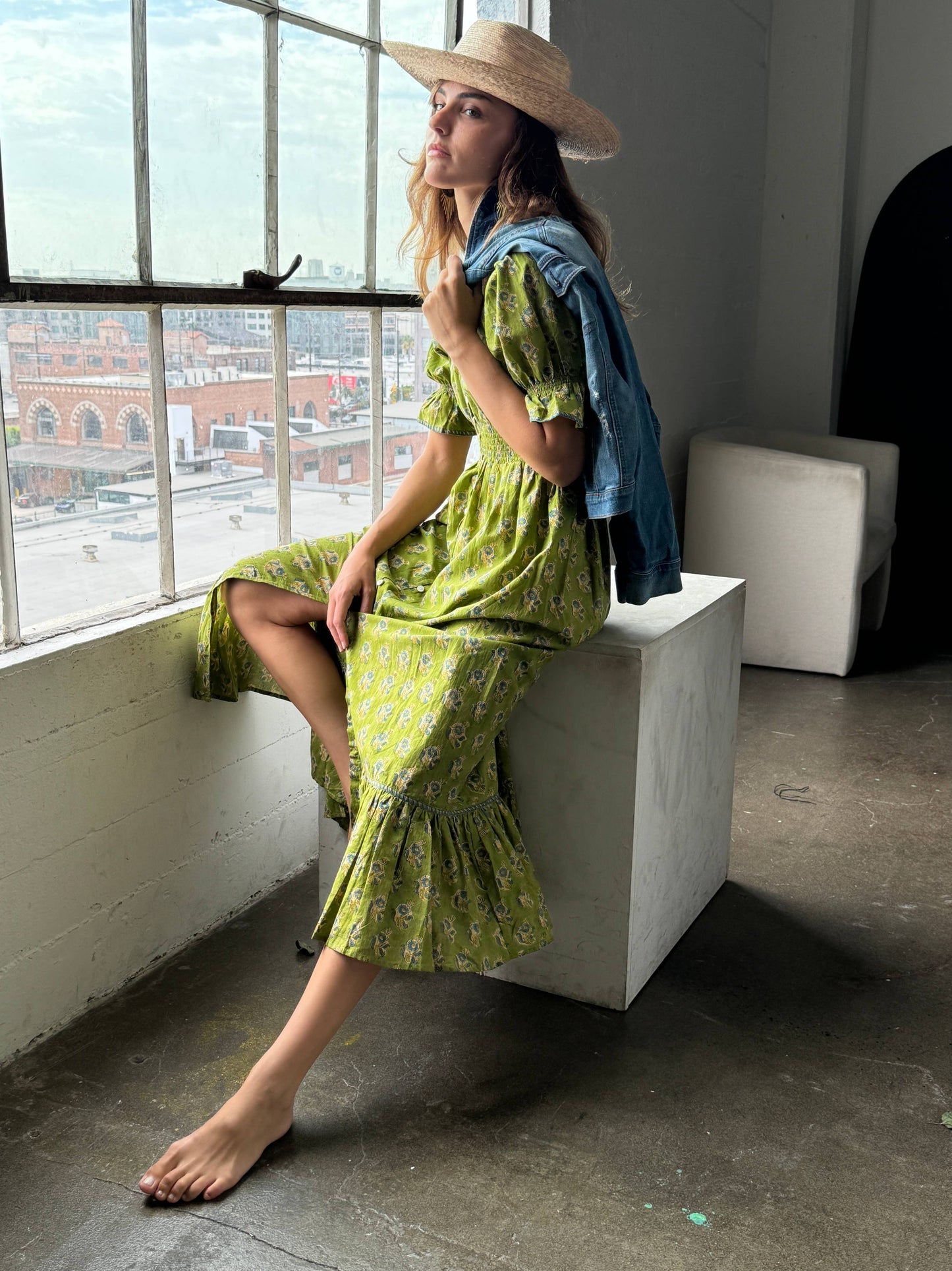 Woman in a green dress and straw hat sitting by a window in an urban setting.