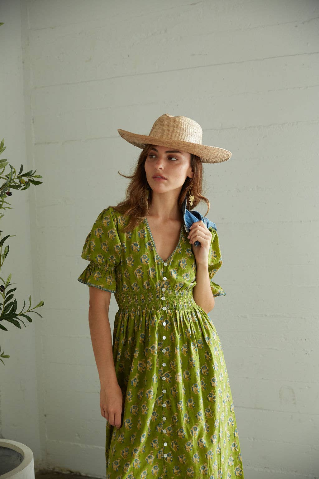 Woman wearing a green floral dress and straw hat holding a blue bag against a white wall.