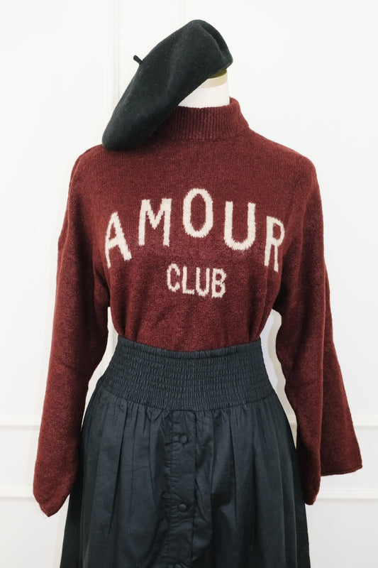 A dark red knit sweater with 'AMOUR CLUB' knitted in cream across the chest, styled on a mannequin with a black beret and a black skirt against a white paneled wall.