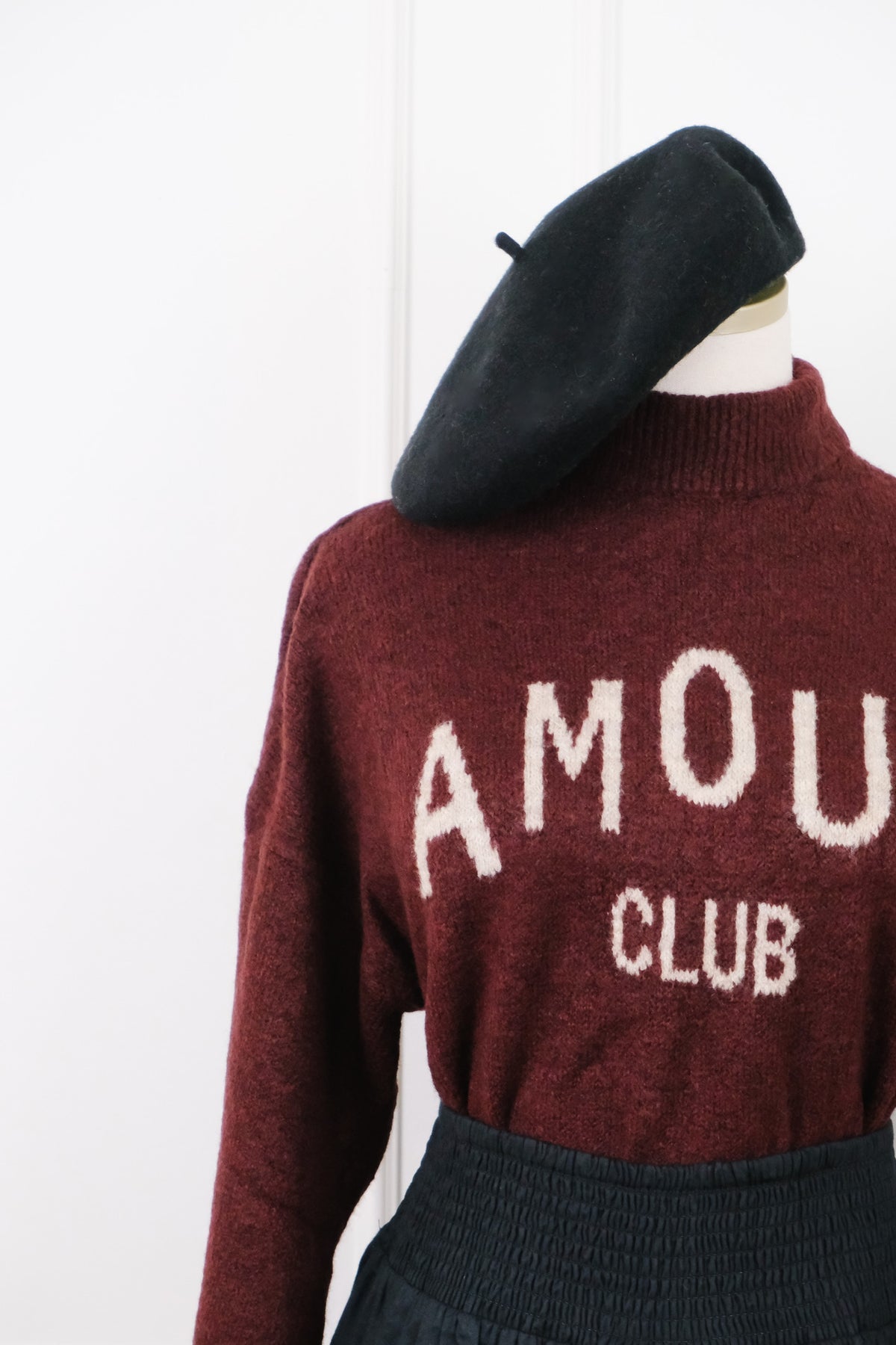 A burgundy knit sweater with 'AMOUR CLUB' knitted in cream on the front, styled on a mannequin with a black beret and dark skirt against a white background.