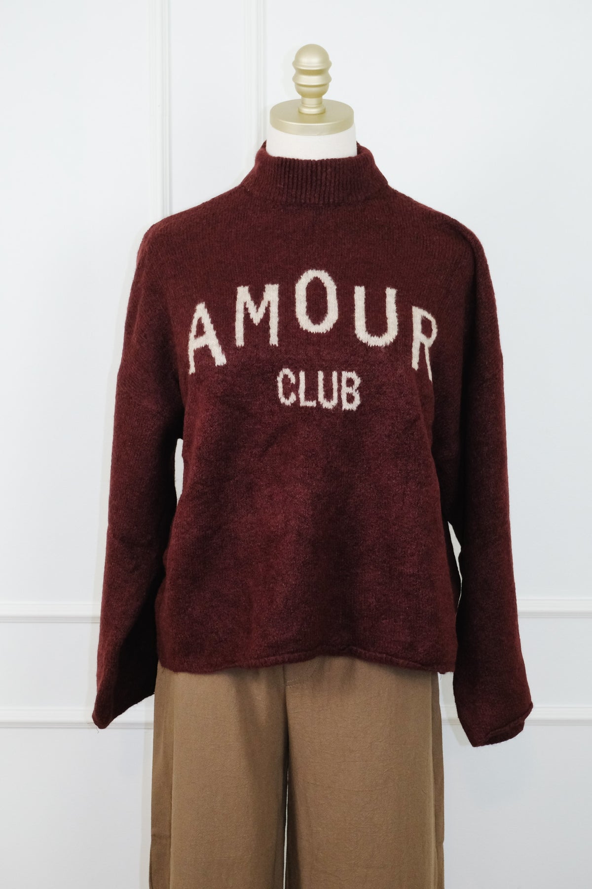 A maroon knit mock-neck sweater with 'AMOUR CLUB' knitted in cream across the chest, displayed on a mannequin torso with tan trousers against a white paneled wall.