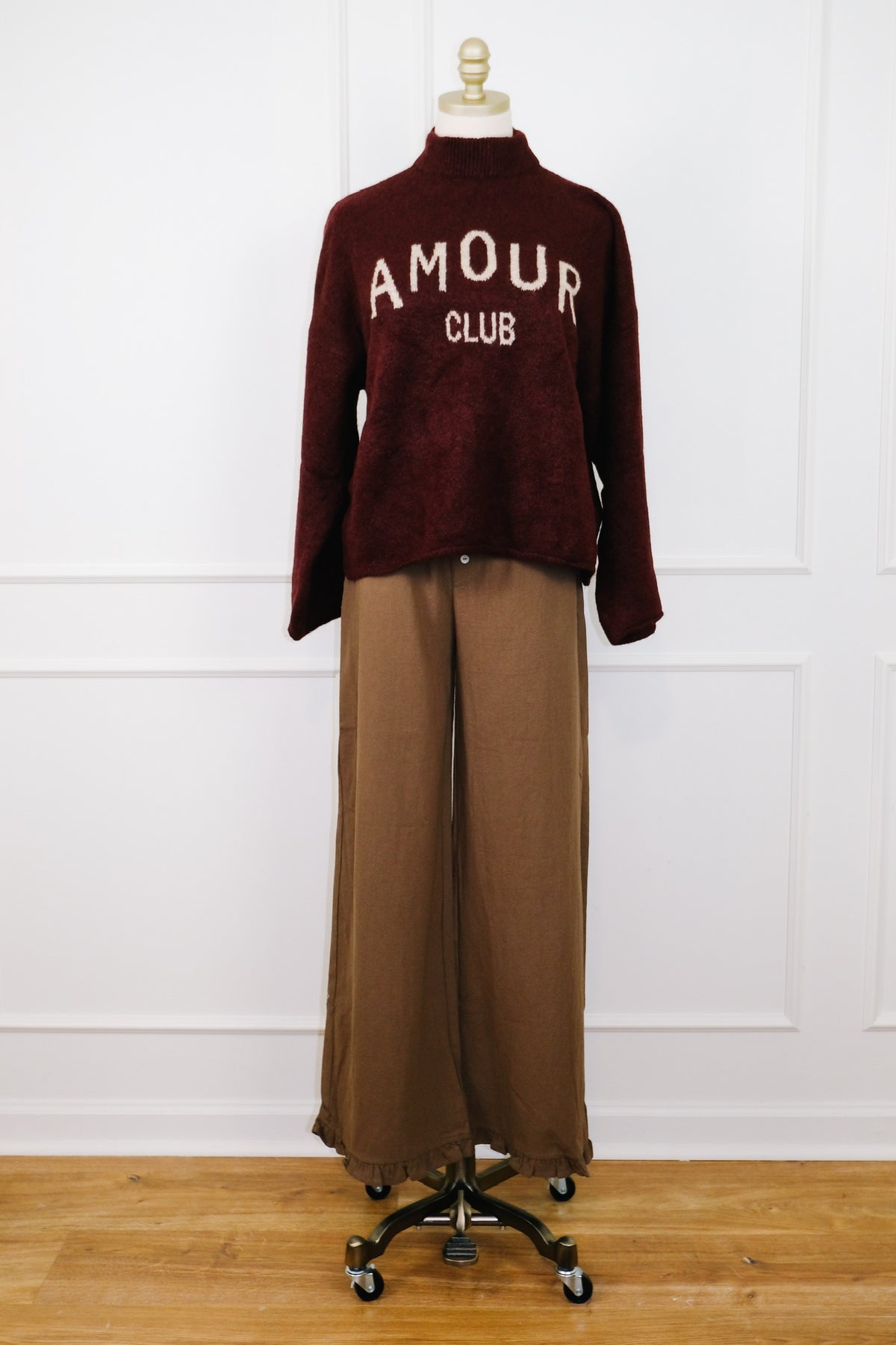 A two-piece outfit displayed on a mannequin, featuring a dark burgundy mock-neck knit sweater with 'AMOUR CLUB' printed in white across the chest, paired with wide-leg brown trousers, set against a white paneled wall and light wood floor.