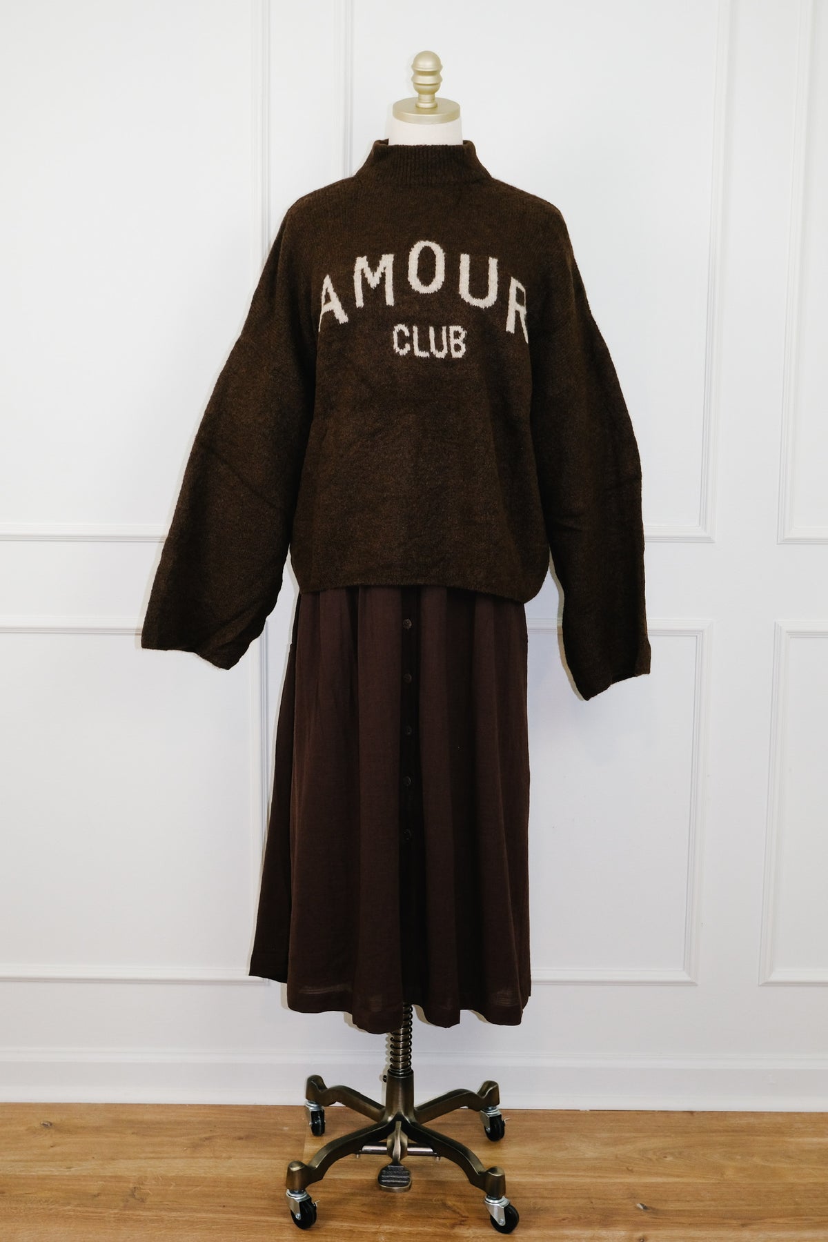 A dark brown cropped knit cardigan with 'AMOUR CLUB' embroidered in white across the chest, displayed on a dress form with a matching dark brown midi skirt against a white paneled wall.