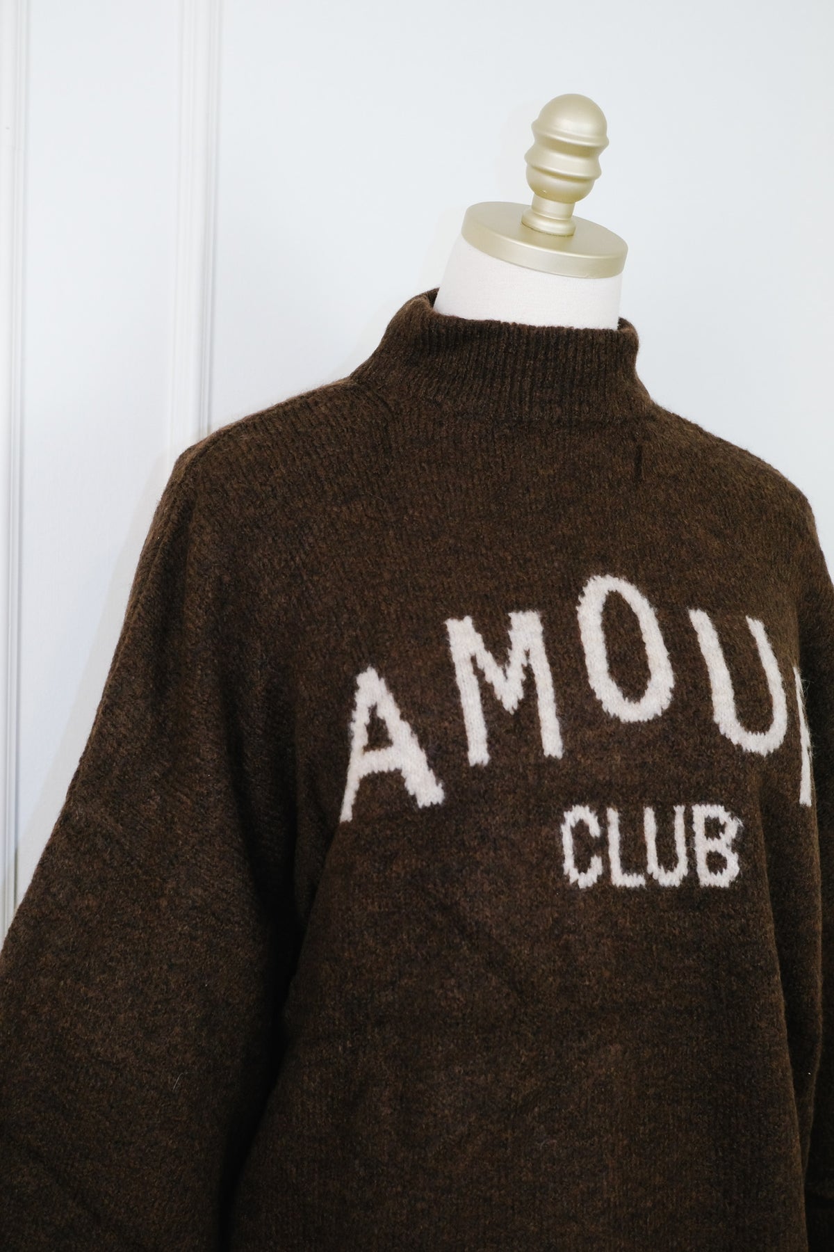 A dark brown knit mock-neck sweater with 'AMOUR CLUB' knitted in white across the chest, displayed on a cream dress form mannequin against a white paneled wall.