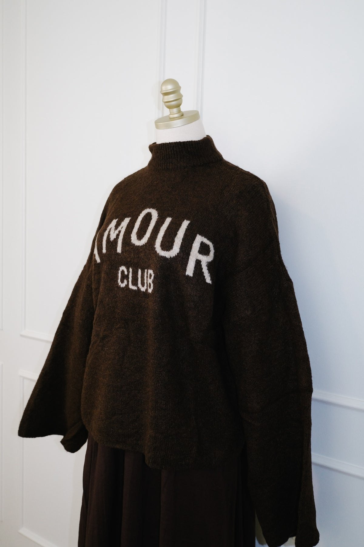 A dark brown fuzzy knit sweater displayed on a mannequin torso against a white paneled wall. The sweater features the words 'AMOUR CLUB' knitted in white across the chest.