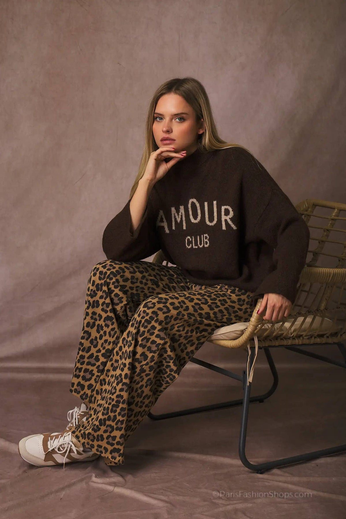 A woman sits on a woven chair against a textured taupe backdrop, wearing a dark brown sweatshirt with 'AMOUR CLUB' printed in white and leopard-print wide-leg pants.