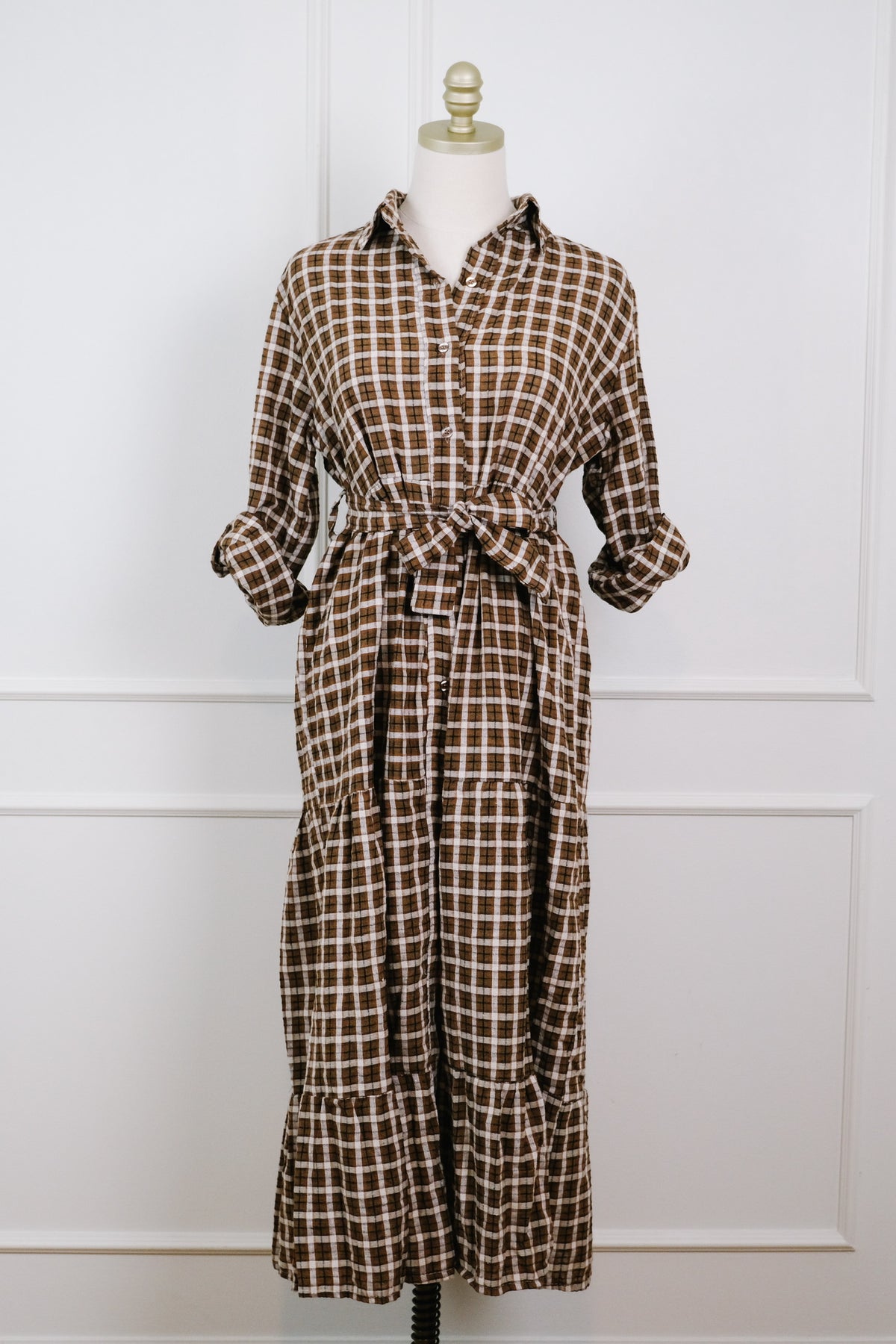 A brown and beige checkered long-sleeve shirt dress displayed on a cream mannequin against a white paneled wall.