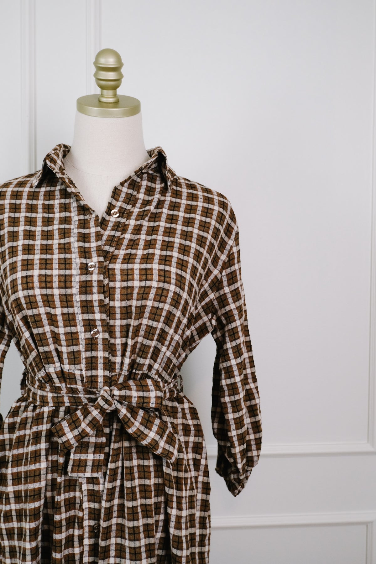 A brown and beige plaid shirt dress displayed on a cream dress form with a gold finial, set against a white paneled wall.