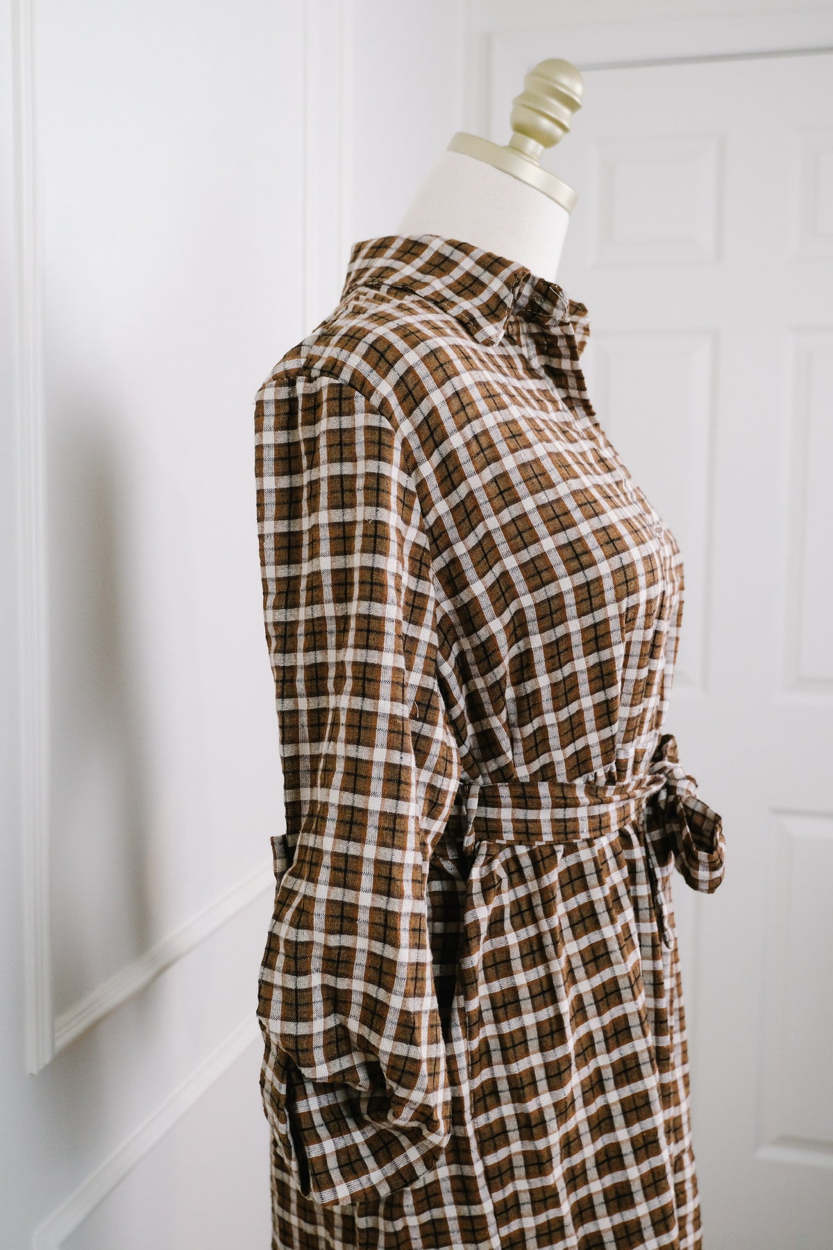 A brown and beige plaid long-sleeve shirt dress displayed on a cream mannequin torso, set against a white paneled wall and door background.