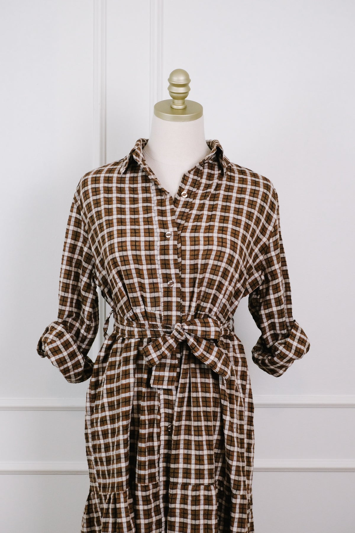 A brown and beige plaid shirt dress displayed on a cream dress form with a gold finial, set against a white paneled wall.