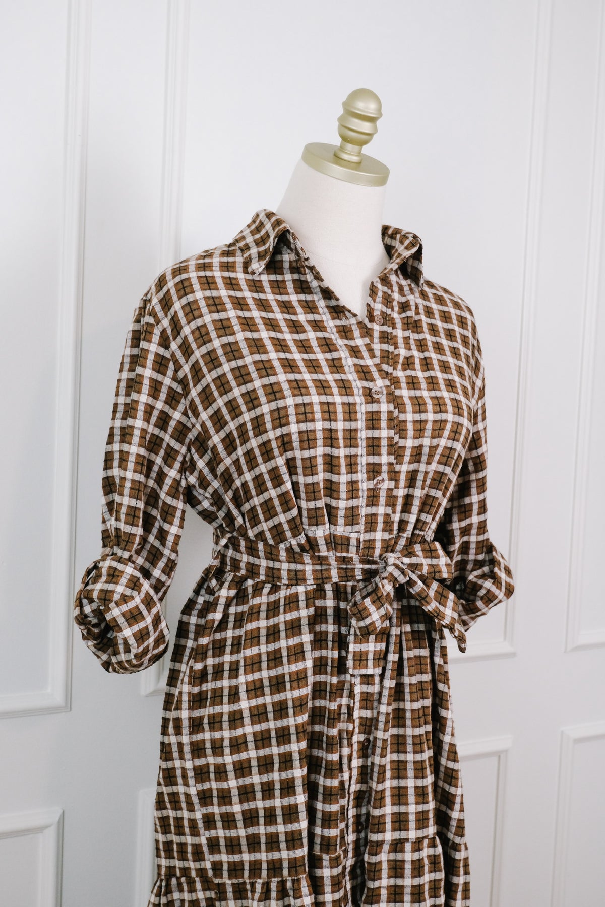 A brown and beige plaid shirt dress displayed on a cream dress form mannequin against a white paneled wall.