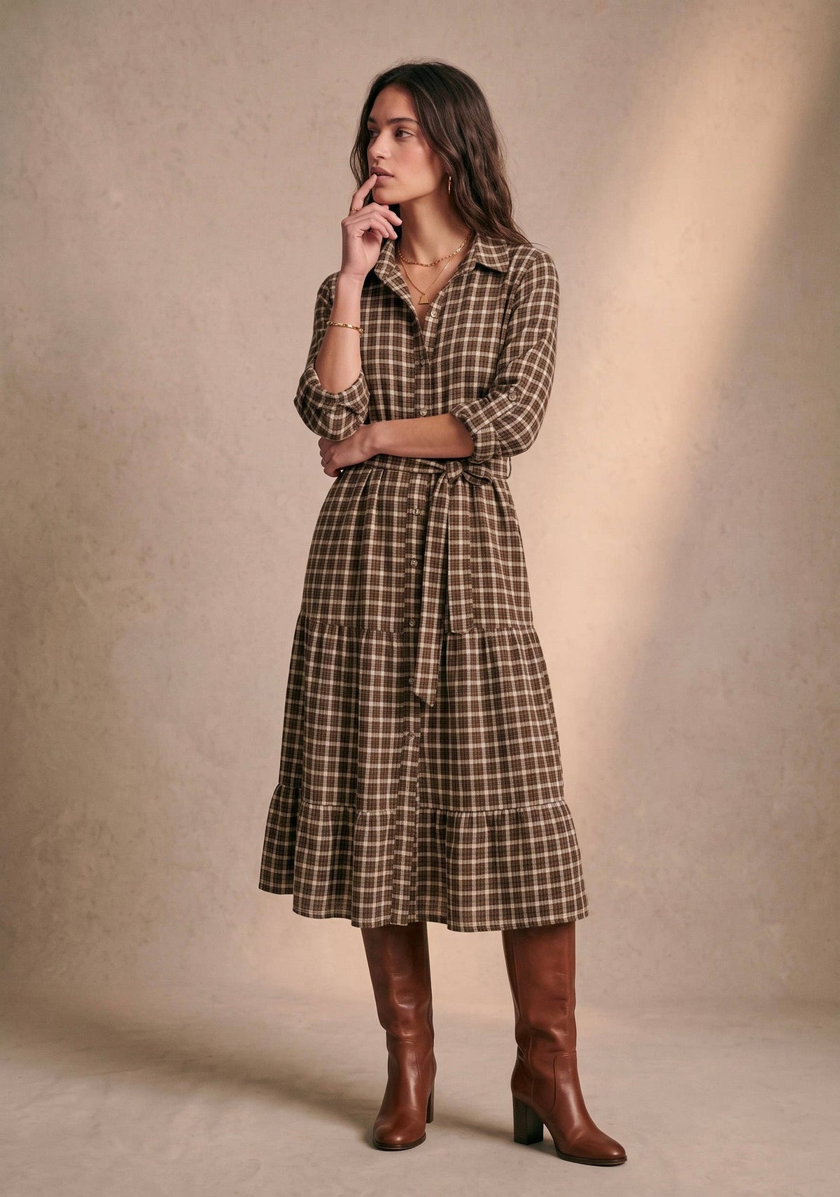 A woman models a brown and beige plaid midi shirt dress with a tie waist, paired with brown knee-high boots, against a textured beige studio backdrop.
