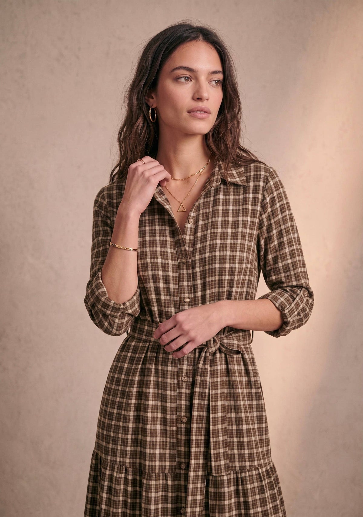 A woman wearing a brown and beige plaid shirt dress with a tie waist, standing against a textured beige background.