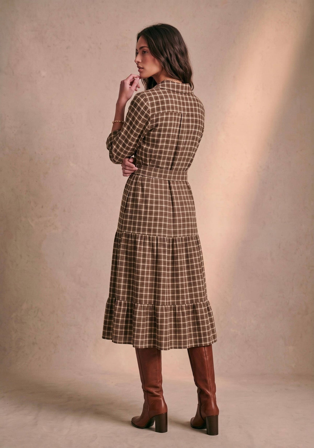A woman wearing a brown and beige plaid midi dress with a tiered skirt and waist tie, paired with brown knee-high boots, photographed from the back against a textured beige studio backdrop.