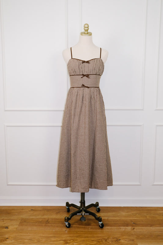 A sleeveless midi dress in a brown and beige plaid pattern, displayed on a cream mannequin dress form over a black rolling stand against a white paneled wall and wooden floor.