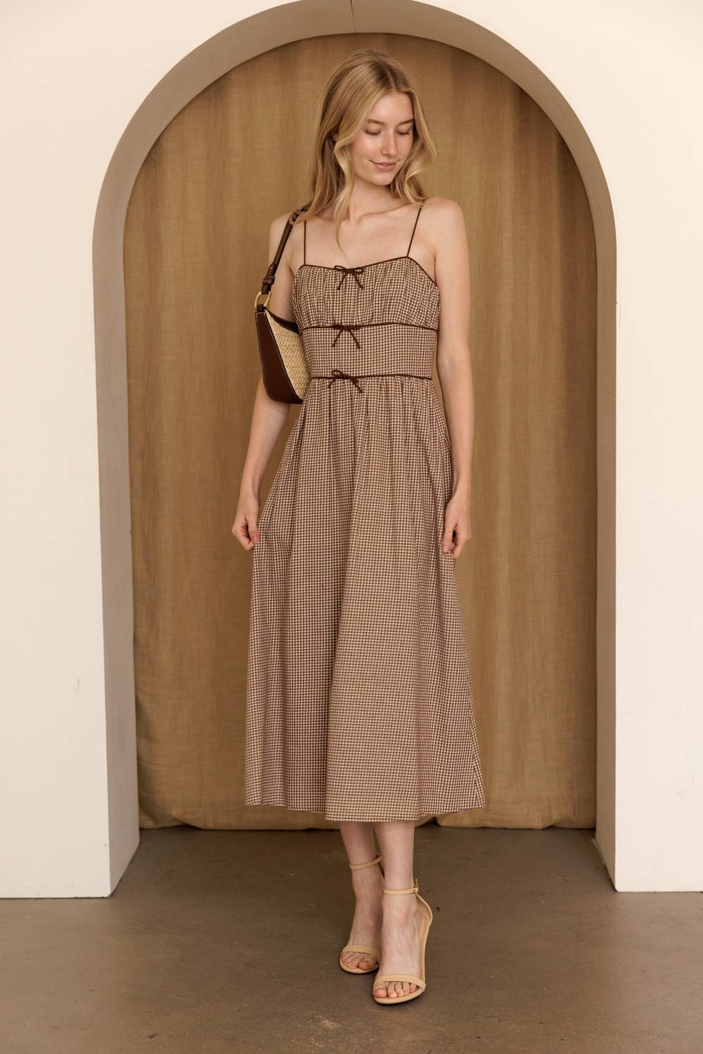A woman modeling a brown and beige gingham midi dress with thin straps and a tie-front detail, standing in front of a wooden arched backdrop.