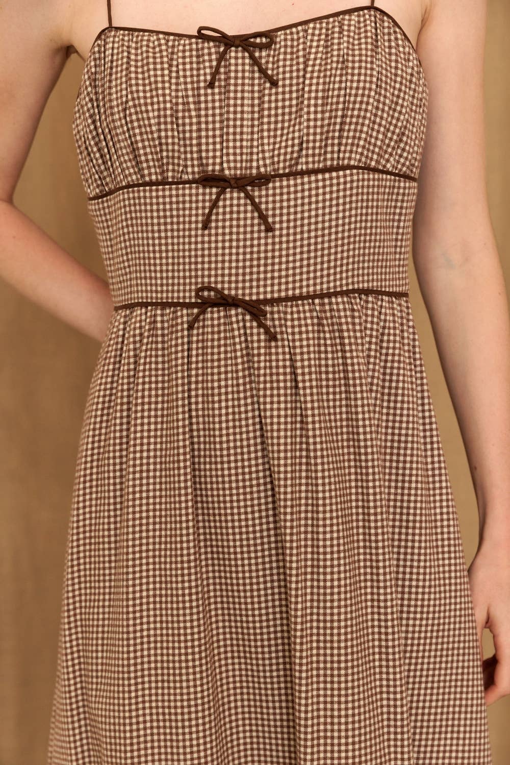 A brown and beige gingham strap dress with thin shoulder straps and three matching waist tie details, shown on a model against a warm beige background.