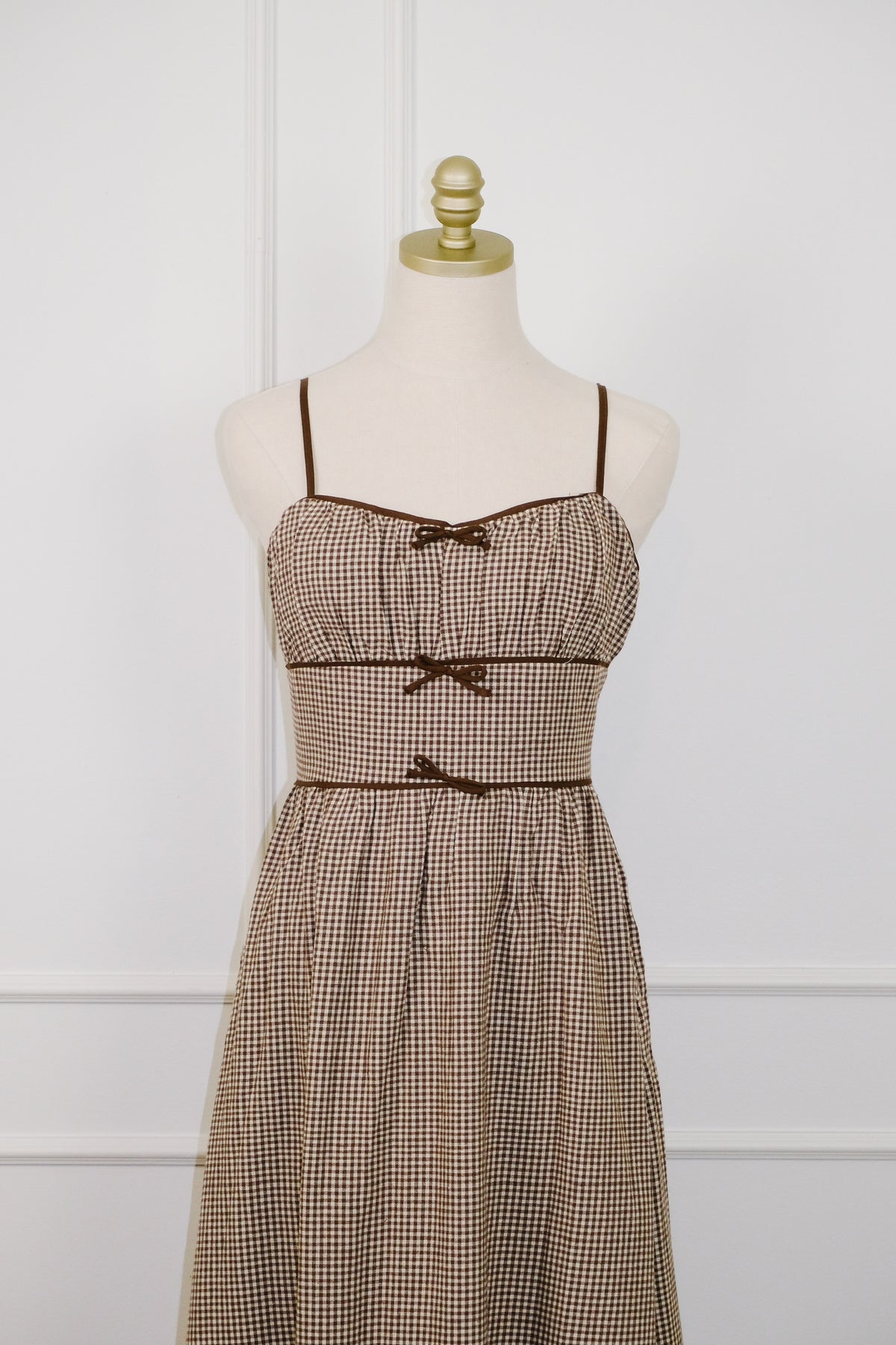 A brown and beige gingham mini dress with thin shoulder straps and three matching fabric bow accents on the bodice, displayed on a cream mannequin with a gold neck cap against a white paneled wall.