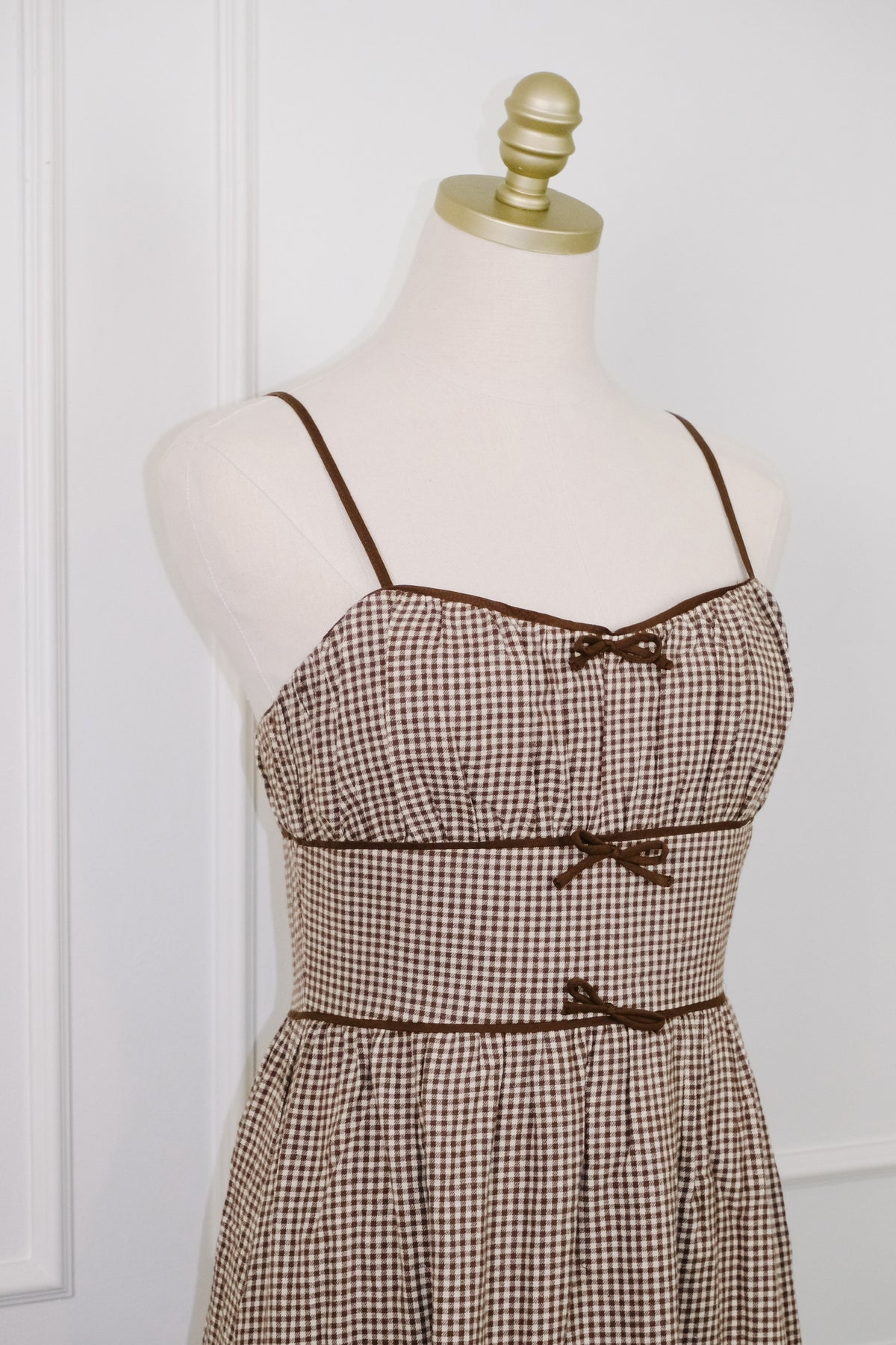A brown and beige gingham check dress displayed on a cream-colored mannequin with a gold neck cap, set against a white paneled wall.