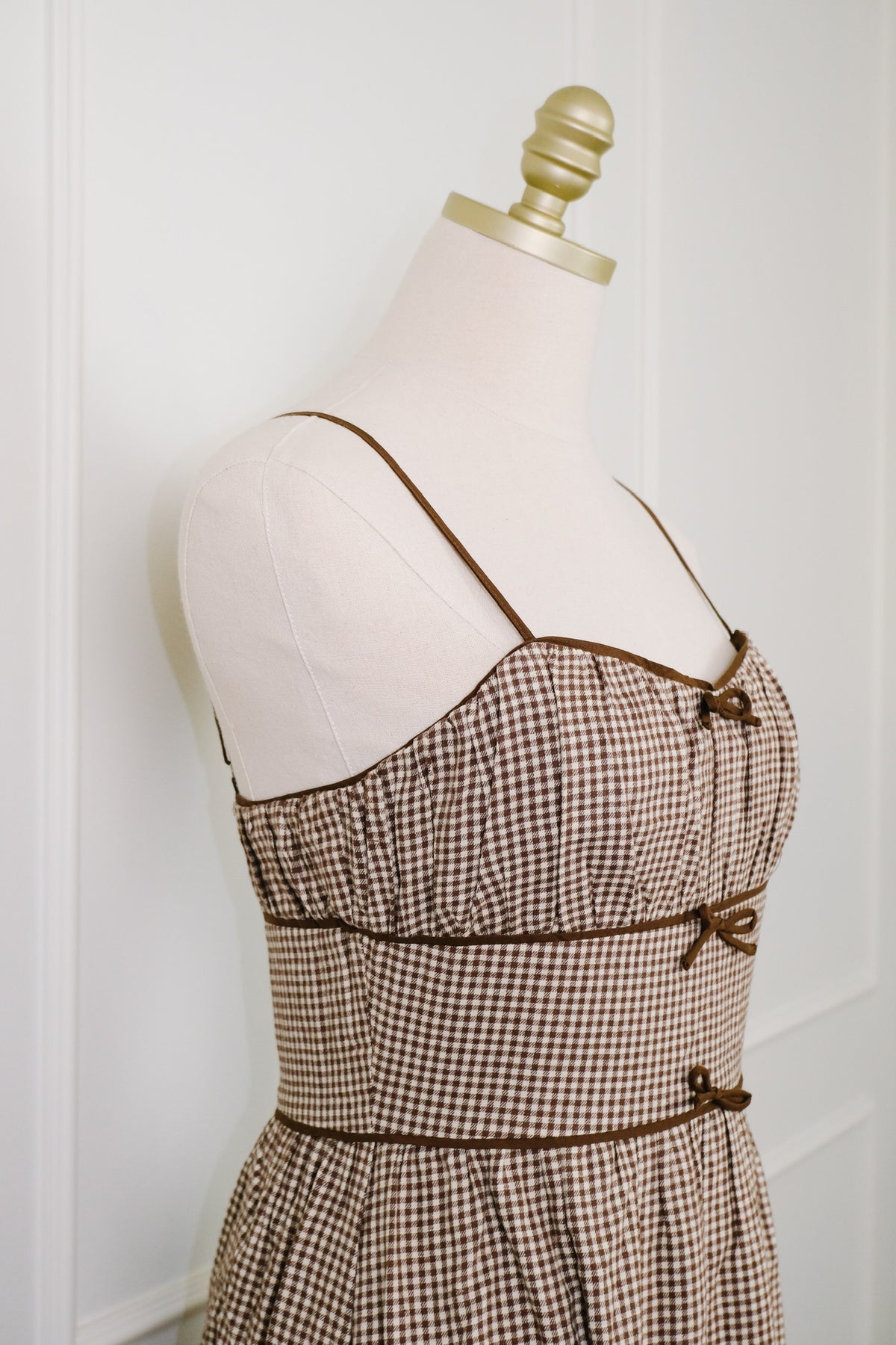 Brown and beige gingham check dress displayed on a cream mannequin torso with a gold neck cap, set against a white paneled wall.