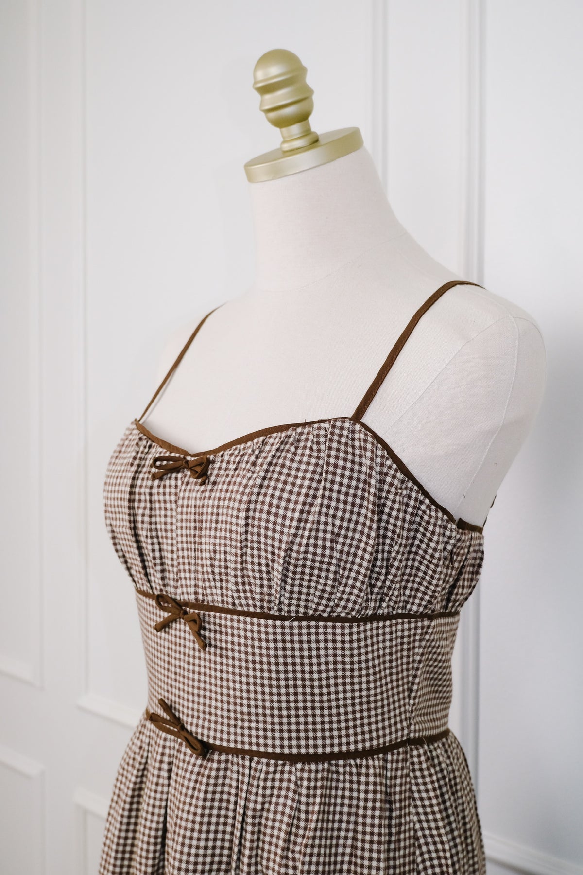Brown and beige gingham check dress displayed on a cream mannequin torso with a gold neck cap, set against a white paneled wall.