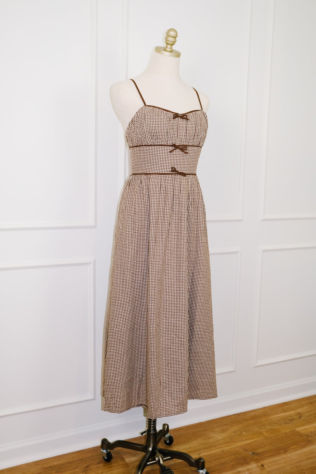 A sleeveless midi dress with a brown and beige gingham pattern, displayed on a cream mannequin dress form over a black rolling stand against a white paneled wall and wood floor.