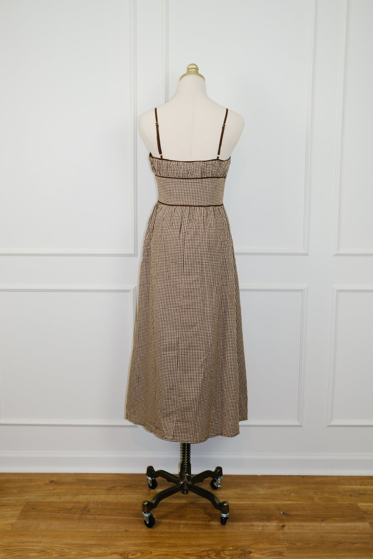 A sleeveless midi dress in a brown and beige textured pattern, displayed on a mannequin dress form over a black rolling stand against a white paneled wall and wooden floor.