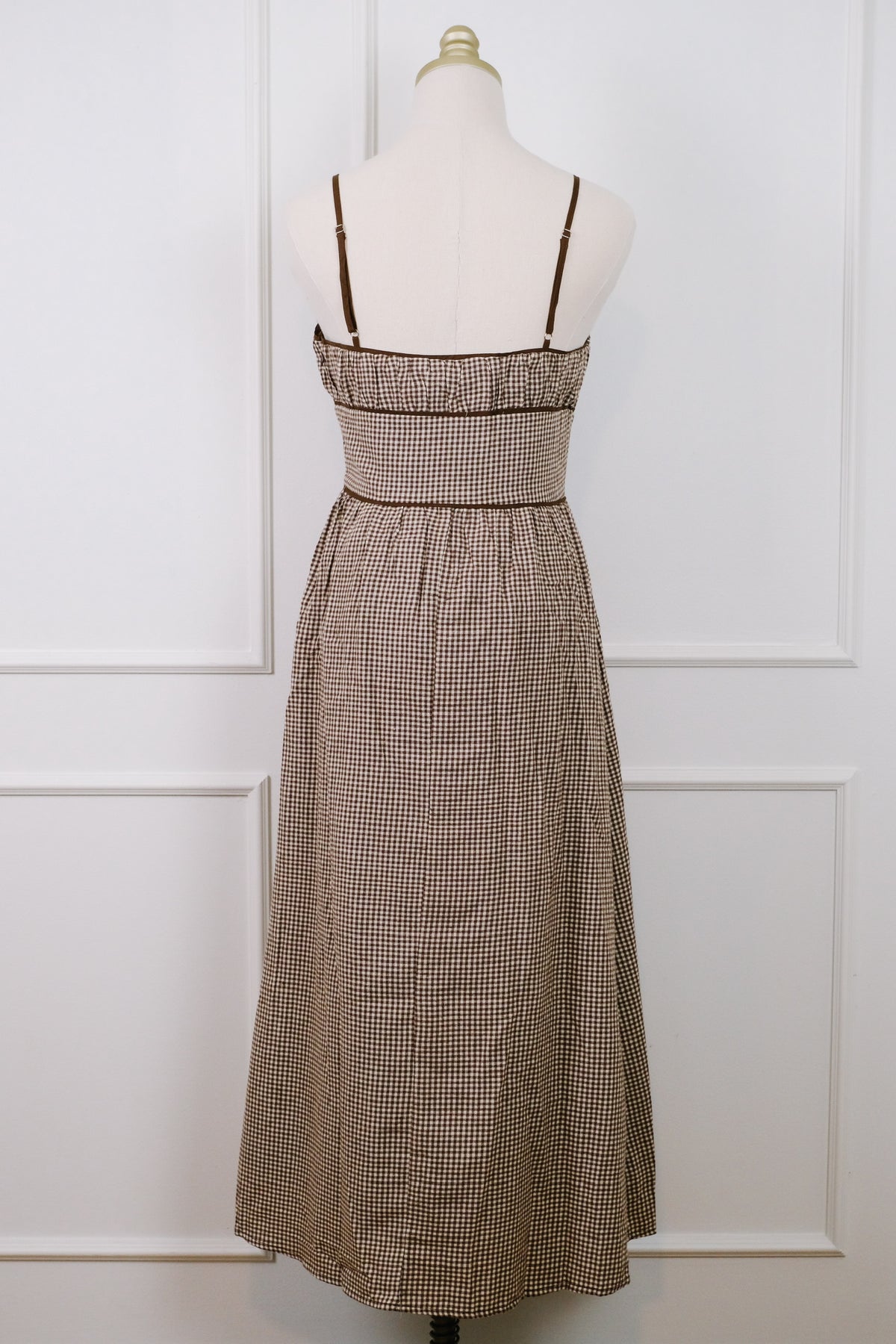 A brown and beige gingham maxi dress displayed on a cream mannequin torso against a white paneled wall. The dress features thin shoulder straps, a ruffled sweetheart neckline, a smocked waist, and a gathered skirt.