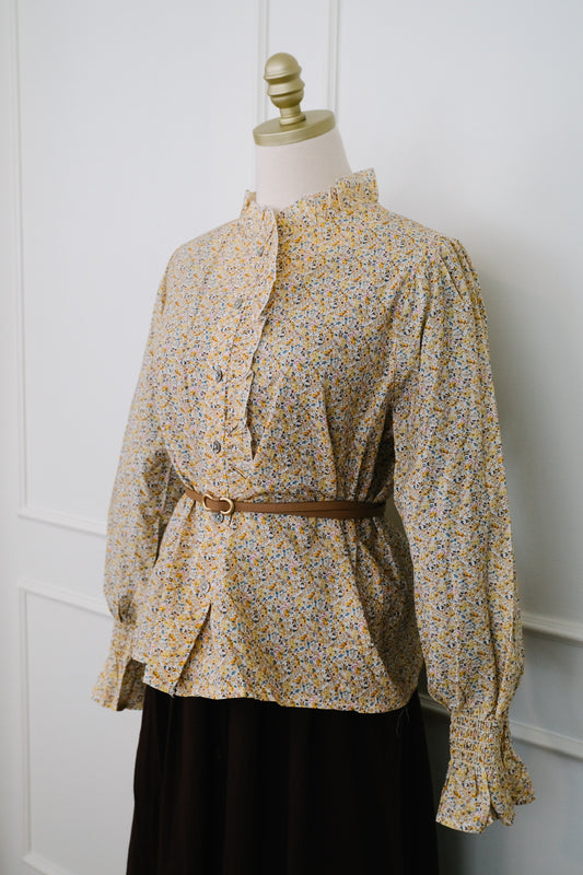 A long-sleeve blouse with a small multicolor floral print on a beige base, styled on a mannequin with a brown belt and a dark skirt against a white paneled wall.
