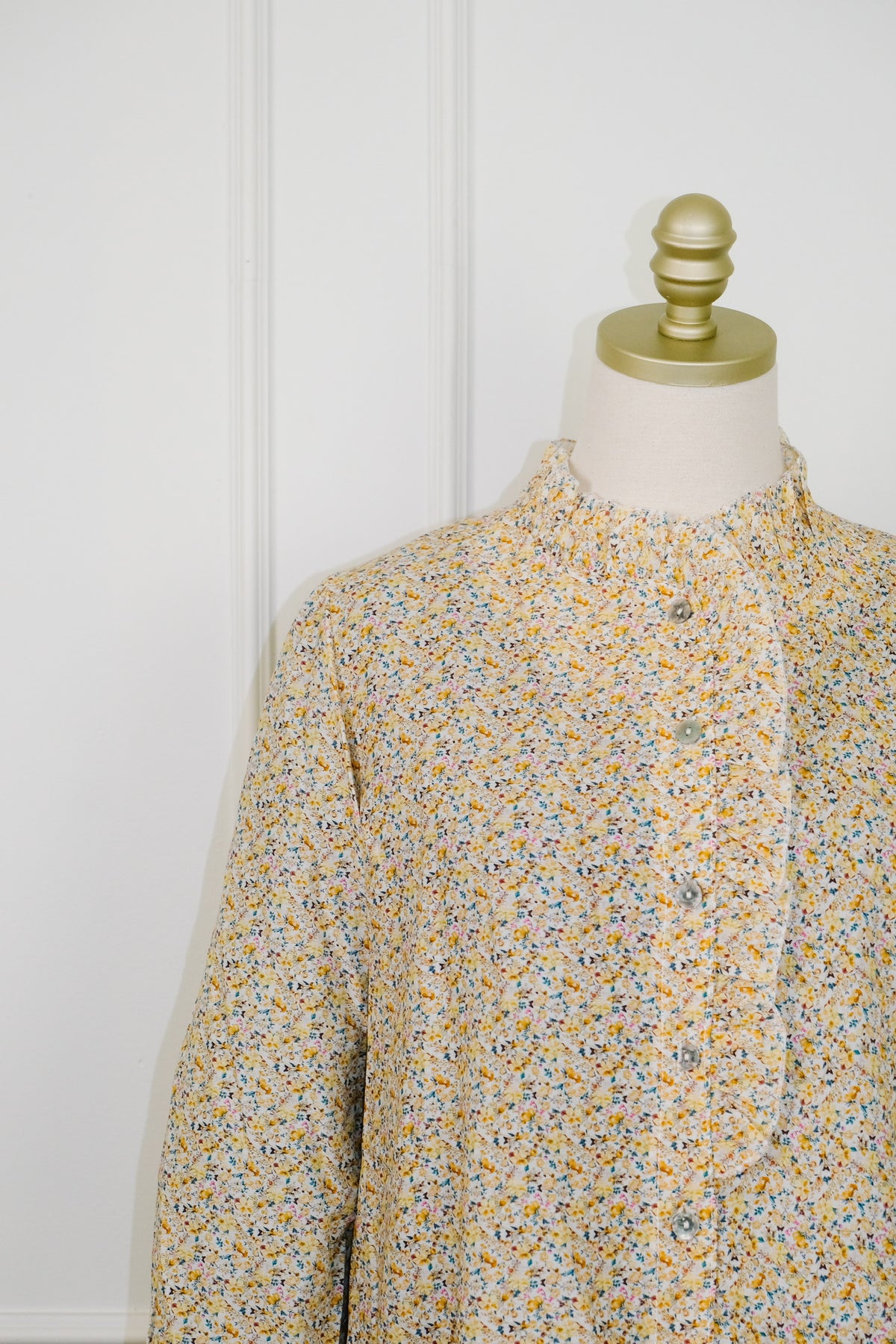 A long-sleeve button-front blouse with a small multicolor floral print on a cream base, displayed on a dress form with a gold finial against a white paneled wall.