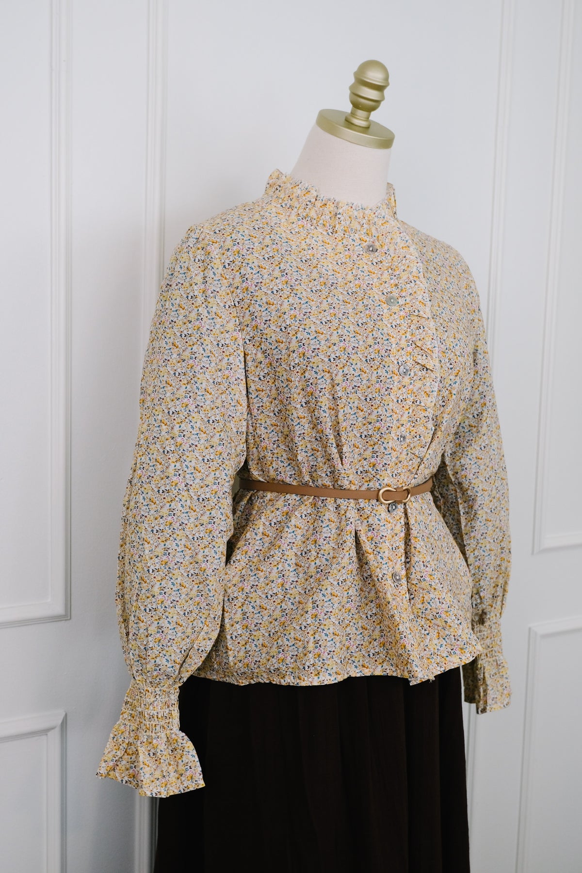 A long-sleeve blouse with a dense small floral print in yellow, blue, and green on a beige base, styled on a dress form with a brown belt and a black skirt against a white paneled door.