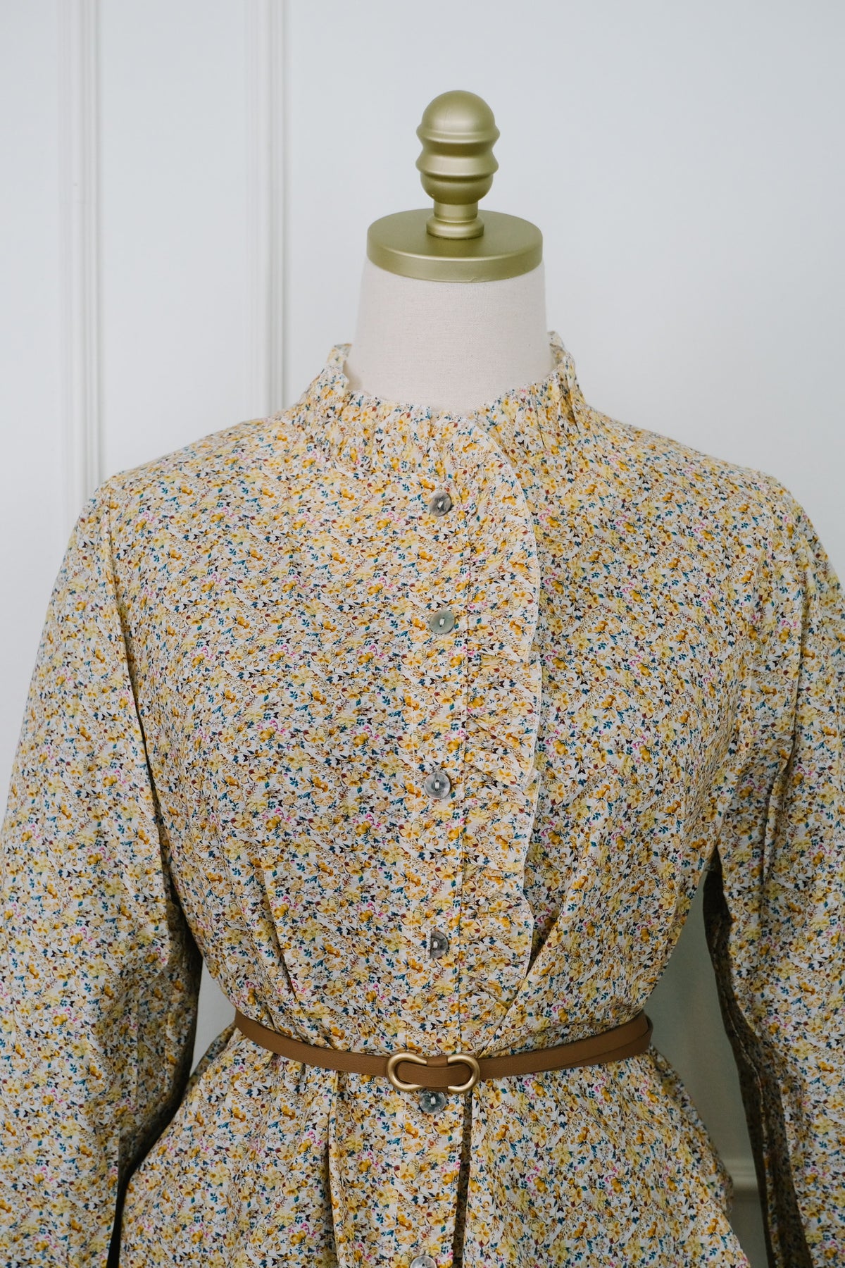 A long-sleeve button-front blouse with a high gathered collar and a small multicolor floral print, displayed on a dress form with a brown belt. The background is a white paneled wall.