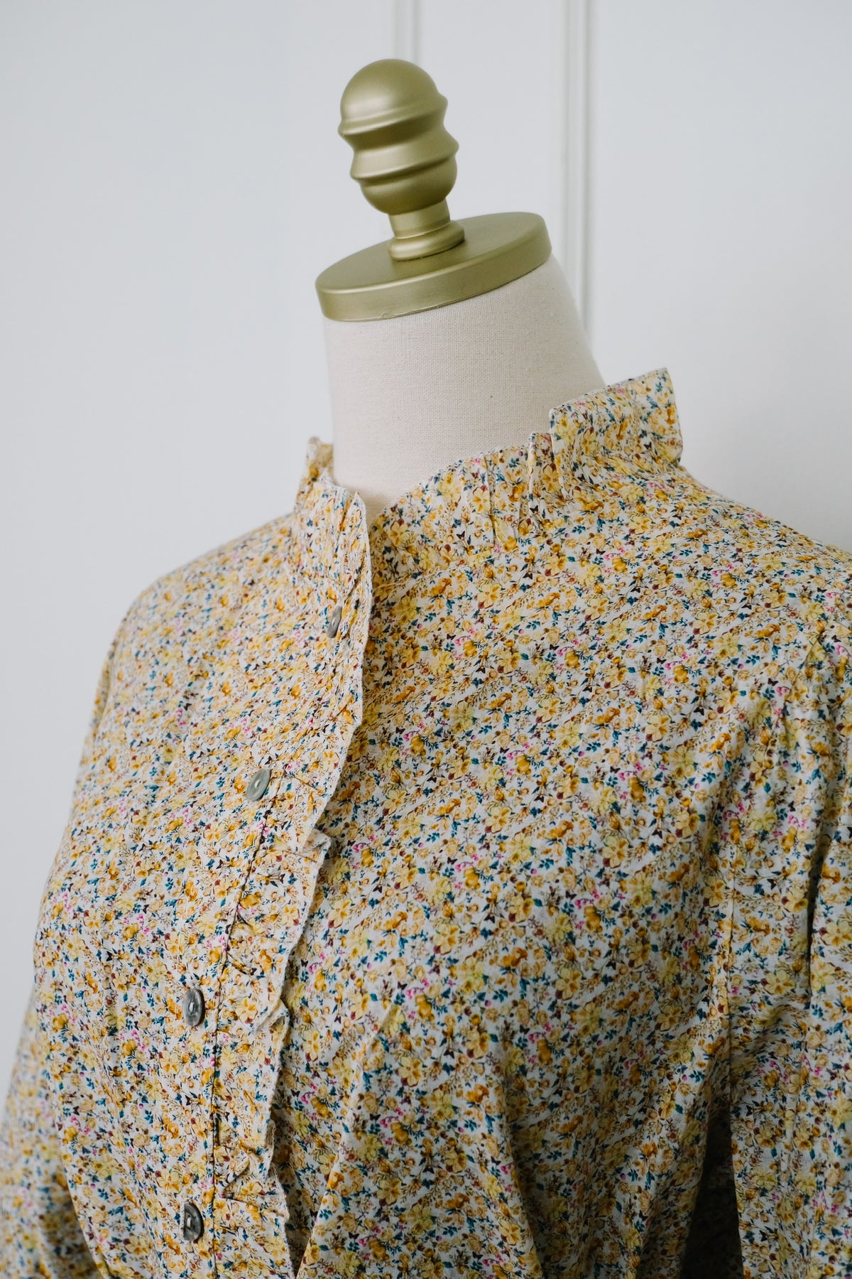 A close-up of a long-sleeve blouse with a ruffled high collar and a dense multicolor floral print, displayed on a cream-colored dress form with a gold finial against a white paneled wall.