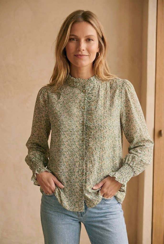 A woman wearing a long-sleeve button-front blouse with a small-scale green and beige floral pattern, paired with light-wash blue jeans, standing against a warm beige wall.