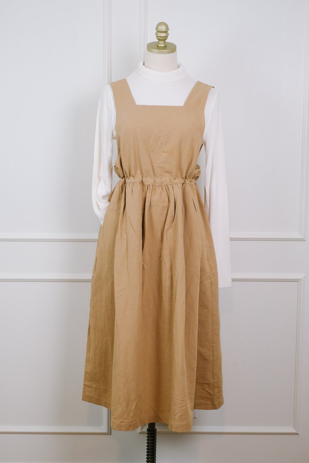 A tan linen pinafore dress styled over a white mock neck long-sleeve shirt, displayed on a dress form against a white paneled wall.