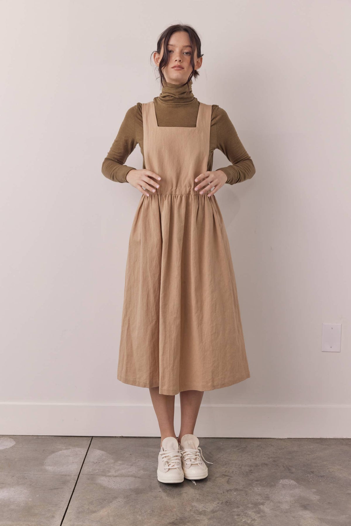 A woman stands indoors wearing a tan linen-look pinafore dress over a taupe turtleneck and white sneakers, posed against a plain off-white wall and concrete floor.