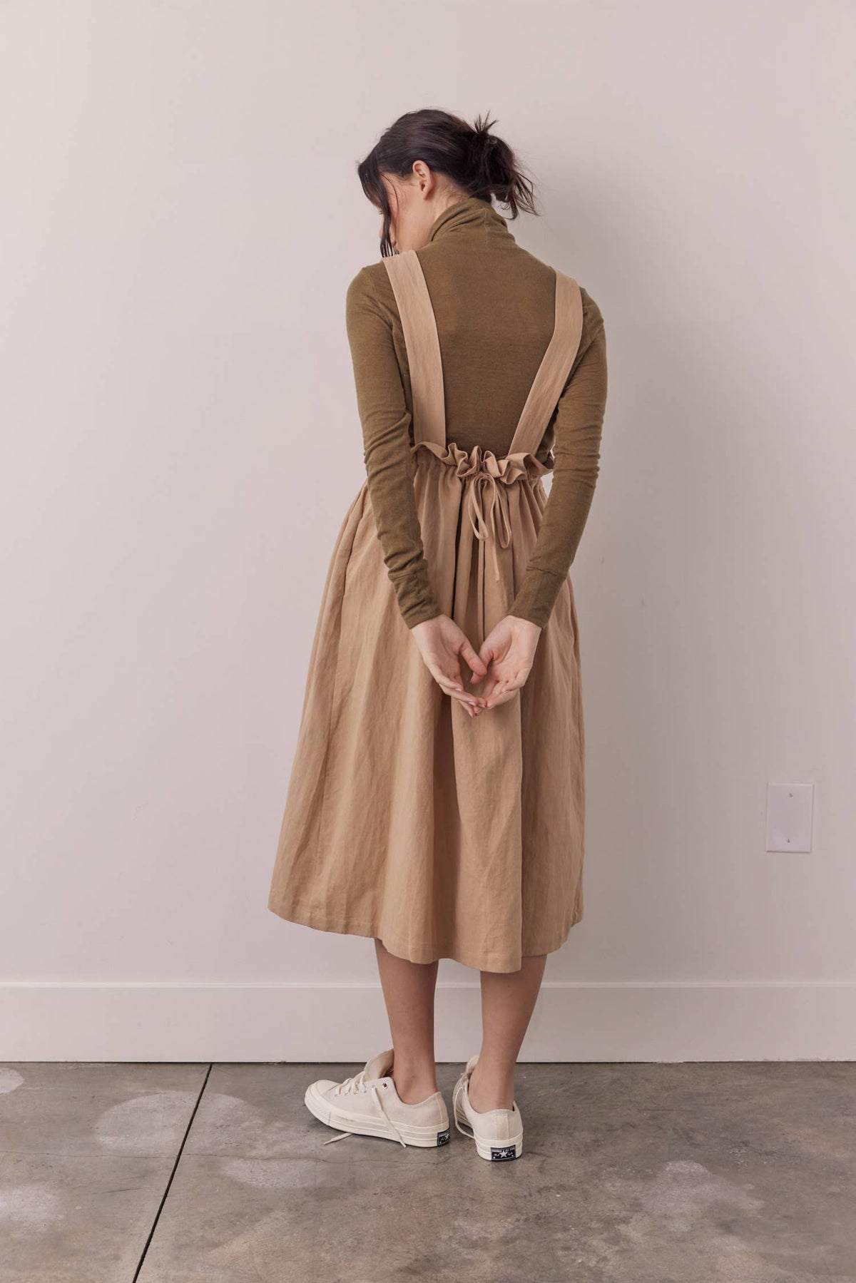 A woman stands with her back to the camera wearing a tan linen-look overall dress over a brown ribbed turtleneck sweater and white sneakers, set against a plain light gray wall and concrete floor.