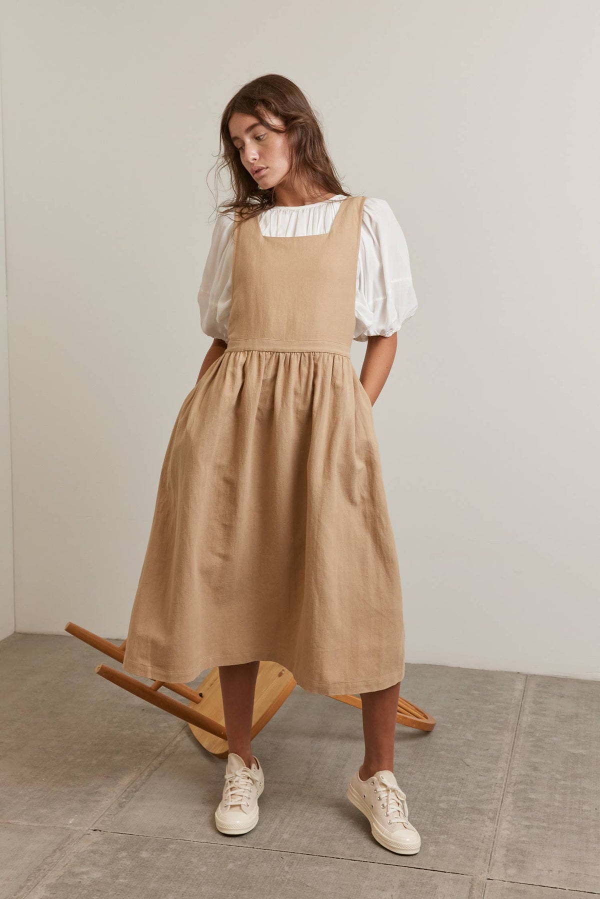 A woman wearing a beige linen pinafore dress over a white puff-sleeve blouse, styled with white sneakers, standing in a minimalist studio setting.