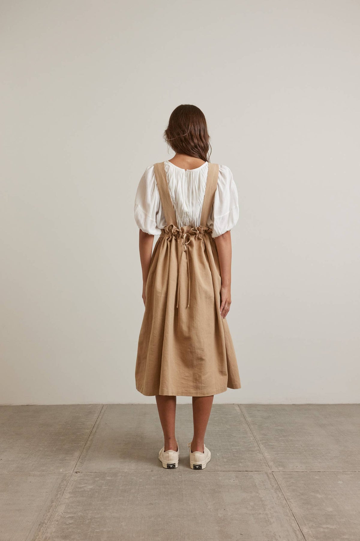 A woman viewed from behind wearing a beige linen-look pinafore dress over a white puff-sleeve blouse, standing on a concrete floor against a neutral beige wall.