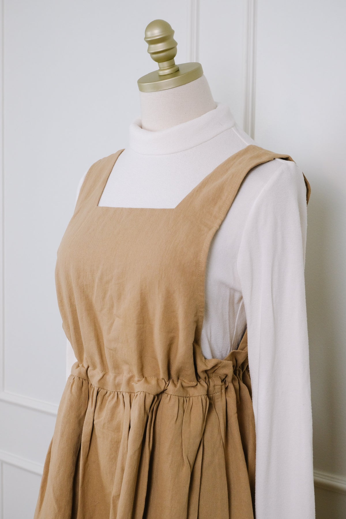 A tan linen pinafore dress styled on a cream dress form mannequin, layered over a white mock neck long-sleeve shirt. The dress features a square neckline, wide shoulder straps, and a gathered elastic waist.