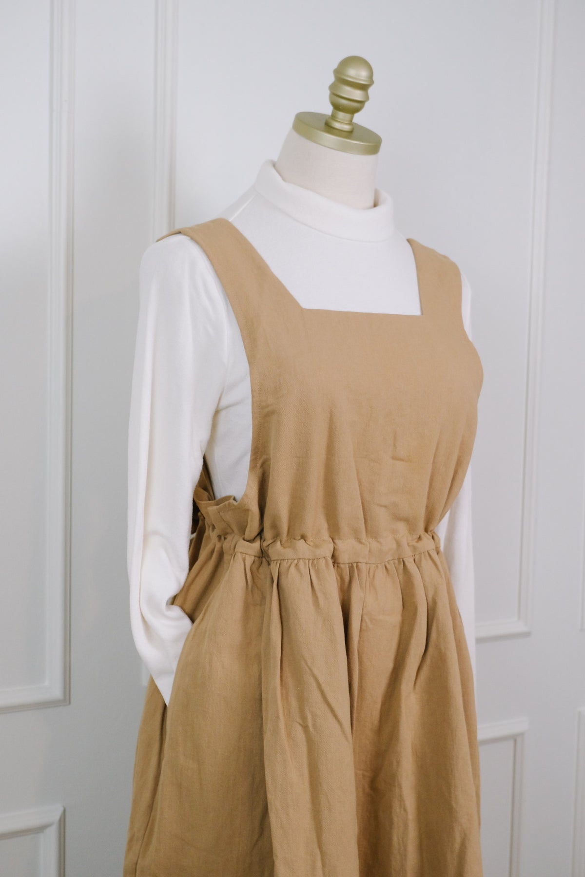A tan linen pinafore dress styled over a white mock neck long-sleeve shirt, displayed on a mannequin dress form against a white paneled door background.