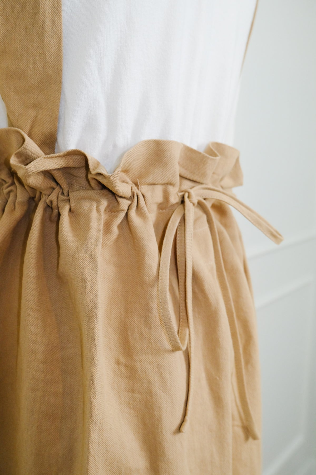 A close-up of a tan linen apron with a gathered paperbag waist and drawstring ties, worn over a white top on a mannequin.