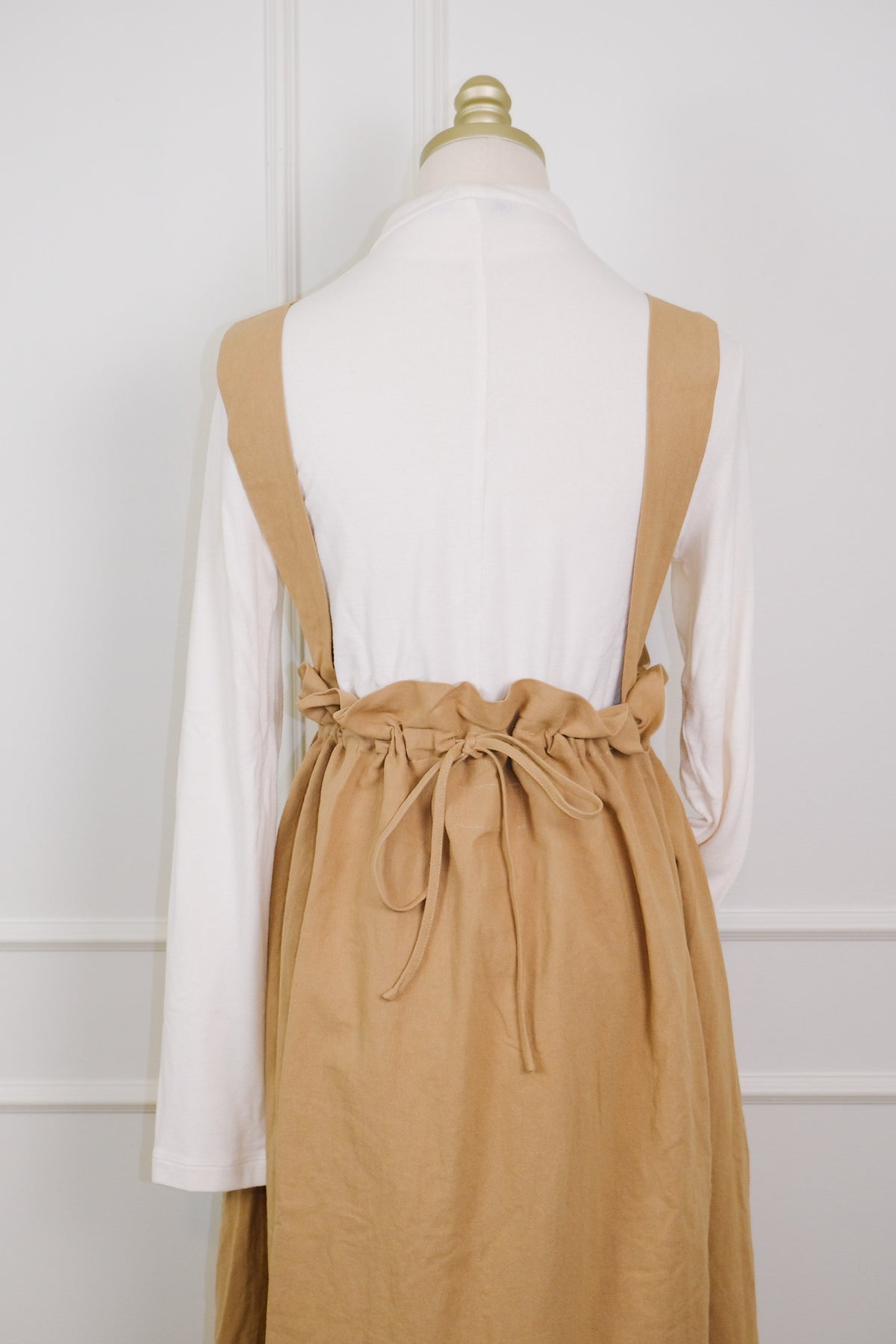 A tan linen pinafore apron with adjustable shoulder straps and a paperbag waist tie, styled over a white long-sleeve shirt on a dress form.