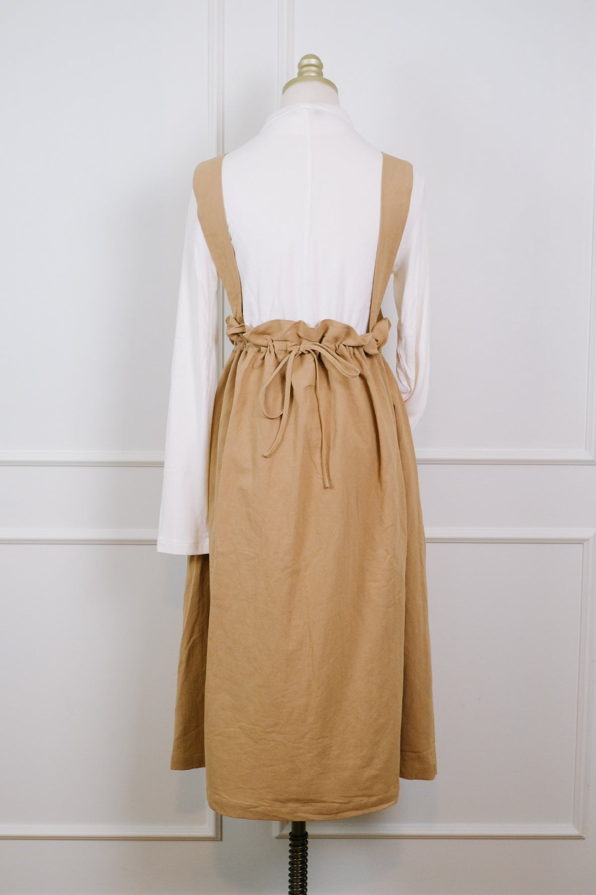 A tan linen bib-style apron or pinafore dress with adjustable shoulder straps and a drawstring waist, layered over a white long-sleeve top and displayed on a dress form against a white paneled wall.