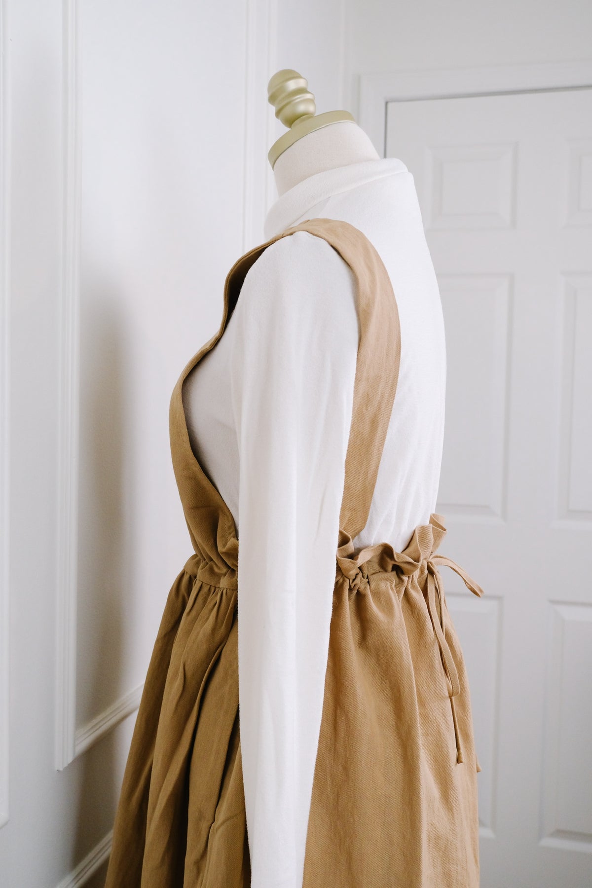 A tan linen pinafore dress styled over a white long-sleeve top on a mannequin, set against a clean white paneled wall and door background.