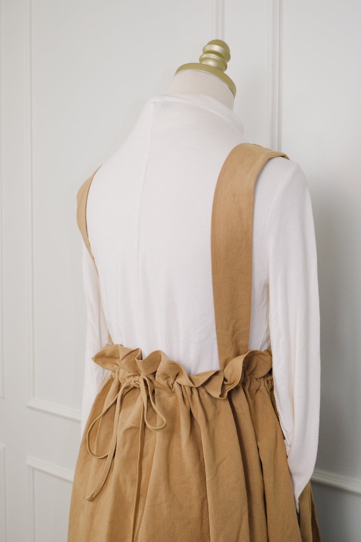 A tan linen pinafore apron with adjustable shoulder straps and a gathered drawstring waist, styled on a white long-sleeve top displayed on a dress form.